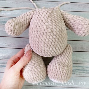 May include: A beige crocheted plush toy with long arms and legs. The toy is being held by a hand, and the bottom of the legs are white.