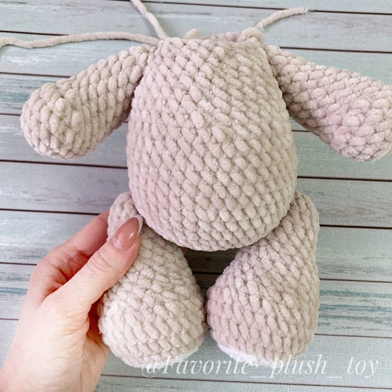 May include: A beige crocheted plush toy with long arms and legs. The toy is being held by a hand, and the bottom of the legs are white.