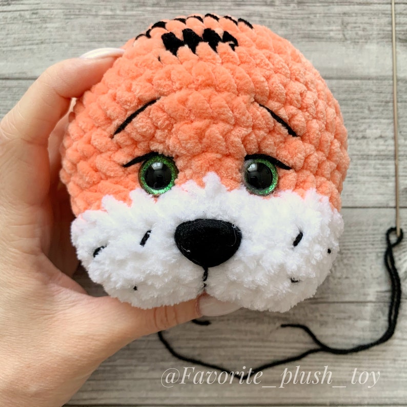 May include: Close-up of a crocheted tiger head with orange fur, white muzzle, black nose, and green eyes. The tiger head is held in a hand.