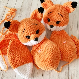 Op de afbeelding: Twee oranje gehaakte vos knuffels met witte accenten. De vossen hebben zwarte ogen en neuzen. De grotere vos heeft een lange, gekrulde staart.