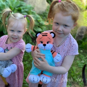 May include: Two young girls are holding a crocheted orange and white tiger plush toy. The tiger is wearing a blue dress. The girls are wearing pink dresses with white polka dots.