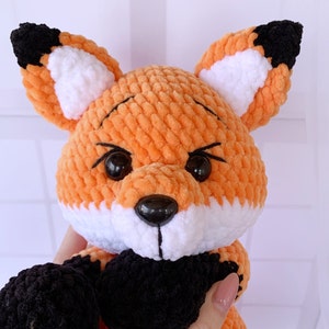 Op de afbeelding: Een gehaakt knuffeldier in de vorm van een vos met oranje, wit en zwart bont. De vos heeft zwarte ogen en een zwarte neus.