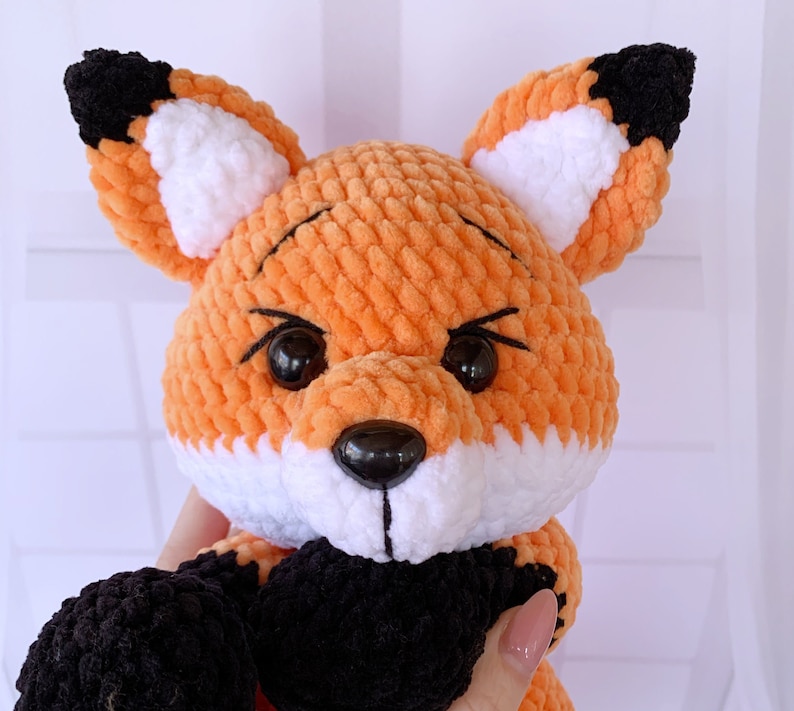 Op de afbeelding: Een gehaakt knuffeldier in de vorm van een vos met oranje, wit en zwart bont. De vos heeft zwarte ogen en een zwarte neus.
