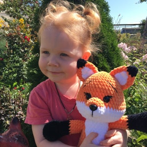 Op de afbeelding: Een jong meisje met rood haar houdt een gehaakt oranje en wit pluche vosje vast. De vos heeft zwarte ogen en een zwarte neus. Het meisje draagt een roze shirt en heeft haar haar in staartjes.