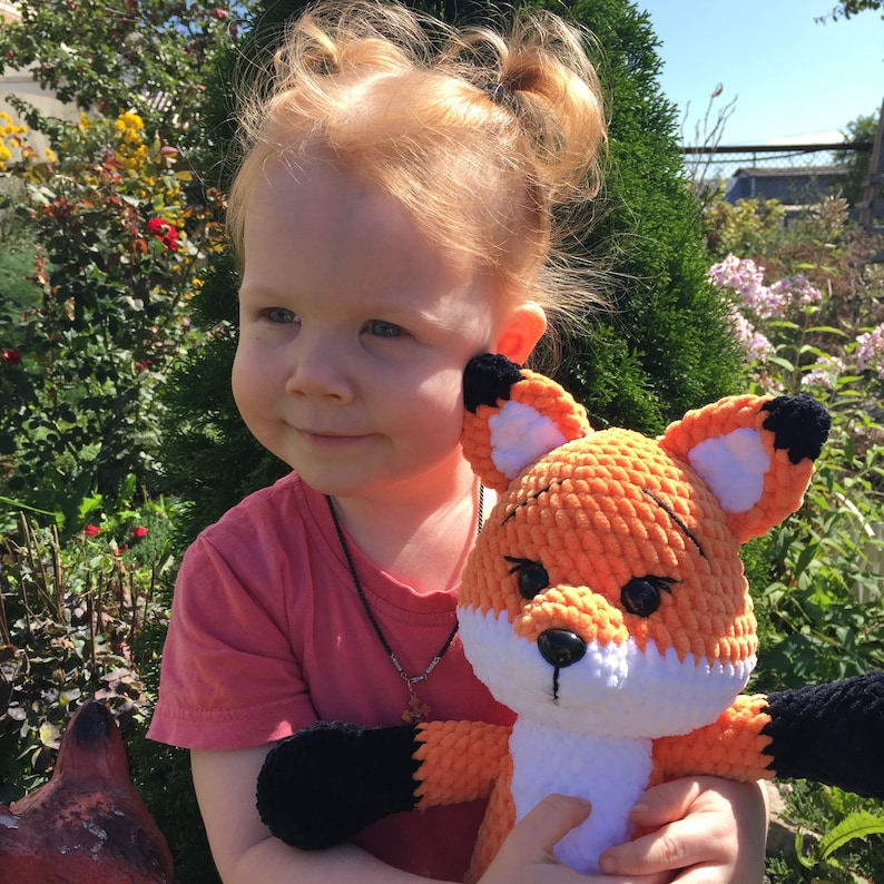 Op de afbeelding: Een jong meisje met rood haar houdt een gehaakt oranje en wit pluche vosje vast. De vos heeft zwarte ogen en een zwarte neus. Het meisje draagt een roze shirt en heeft haar haar in staartjes.