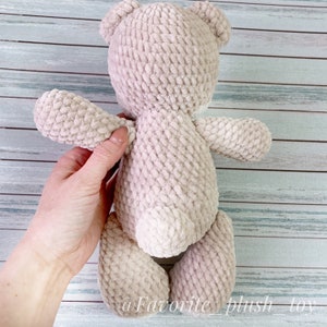 May include: A beige crocheted teddy bear with a fluffy texture. The bear is standing on a wooden surface.
