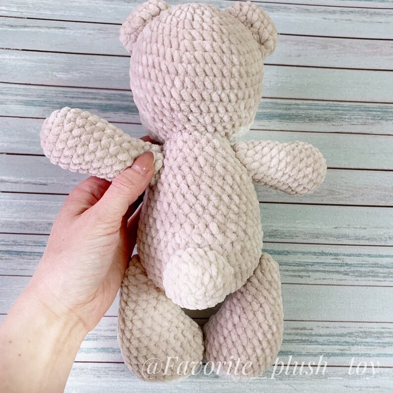 May include: A beige crocheted teddy bear with a fluffy texture. The bear is standing on a wooden surface.