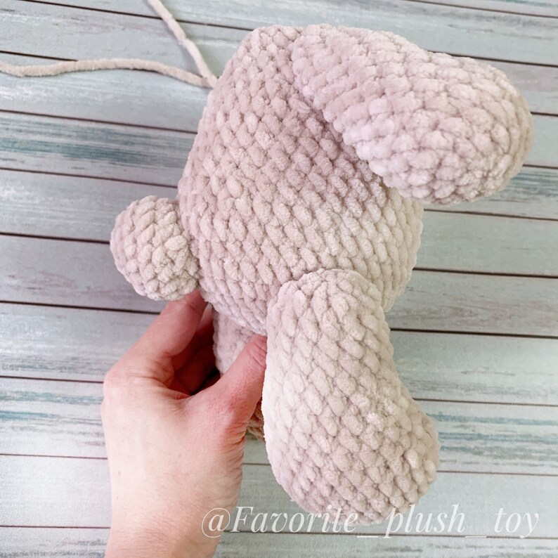 May include: A beige crocheted plush toy in the shape of a bunny rabbit. The toy is being held by a hand, and the back of the toy is facing the camera. The toy has a long, floppy ear and a small, round tail.