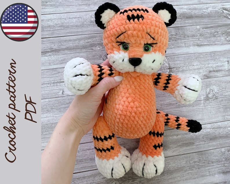 May include: Crocheted orange and black tiger plush toy with white paws and green eyes. The toy is being held by a hand.