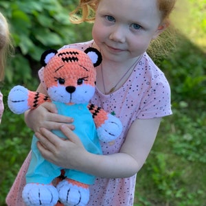 May include: A young girl is holding a crocheted tiger stuffed animal wearing a blue overall. The tiger has orange and black stripes and white fur on its face and paws.