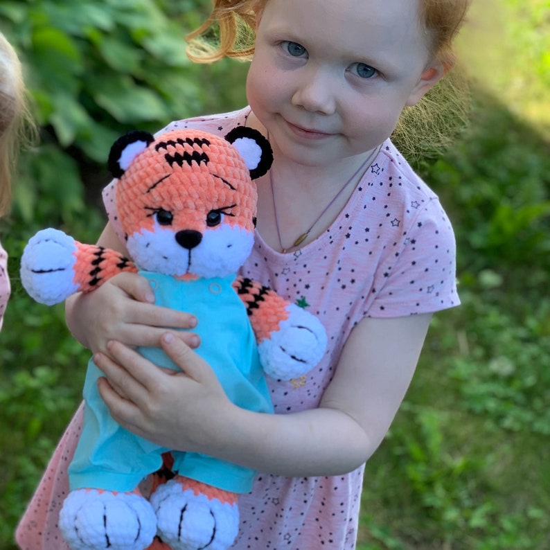May include: A young girl is holding a crocheted tiger stuffed animal wearing a blue overall. The tiger has orange and black stripes and white fur on its face and paws.