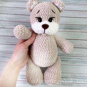 May include: A crocheted plush teddy bear with a beige body, white face, and brown nose. The bear has black eyes and a sad expression.