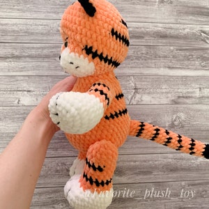 May include: A crocheted orange and black tiger plush toy with white accents. The toy is standing on a wooden surface.