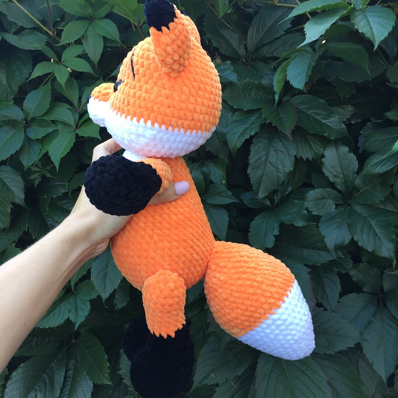 Op de afbeelding: Een gehaakt oranje en wit pluche vosje met zwarte voeten en oren. Het speelgoed wordt vastgehouden door een hand.
