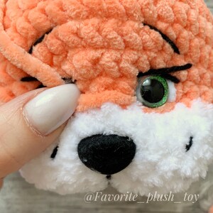 May include: Close-up of a crocheted fox toy with a white and orange fur. The toy has a black nose, green eyes, and black eyelashes. The toy is being held by a hand with white nail polish.