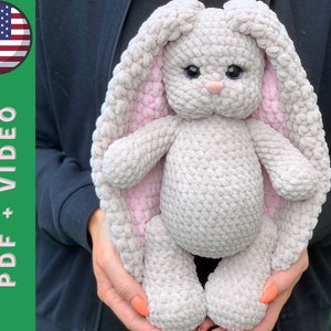 May include: A crocheted plush bunny toy with gray fur and pink ears. The bunny is being held by a person's hands.