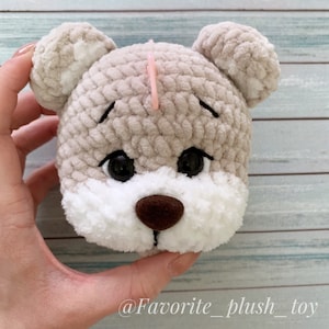 May include: Close-up of a crocheted plush toy bear with brown eyes, a brown nose, and white fur around the mouth. The bear is beige and has a pink stitch marker on the top of its head.