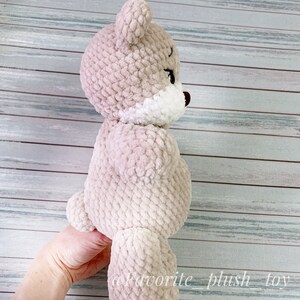 May include: A beige crocheted teddy bear with white accents around the face and a brown nose. The bear is standing on its hind legs with its front paws raised.