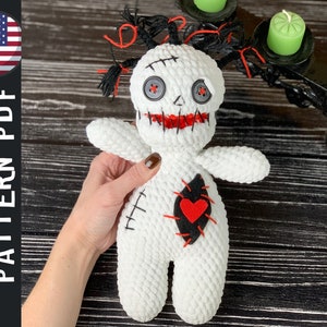May include: White crocheted voodoo doll with red yarn hair, black eyes, and a red heart on its chest. The doll is stitched with black yarn and has a stitched mouth.