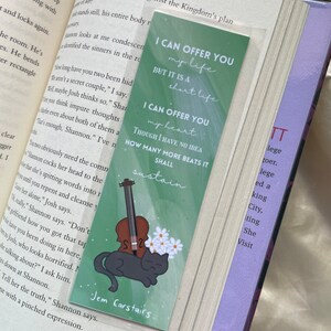 May include: Green bookmark with a black cat and a violin. The bookmark has white flowers and the text "I can offer you my life but it is a short life. I can offer you my heart. Though I have no idea how many more beats it shall sustain." The bookmark also has the text "Jem Carstairs" at the bottom.