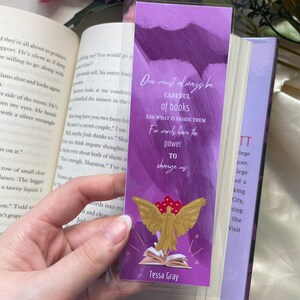 May include: Purple and gold bookmark with a quote about books: "One must always be careful of books and what is inside them, for words have the power to change us." The bookmark features a gold angel with wings spread wide, holding a book open. The text "Tessa Gray" is printed at the bottom of the bookmark.