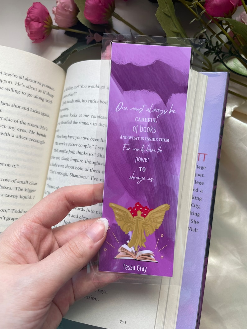 May include: Purple and gold bookmark with a quote about books: "One must always be careful of books and what is inside them, for words have the power to change us." The bookmark features a gold angel with wings spread wide, holding a book open. The text "Tessa Gray" is printed at the bottom of the bookmark.