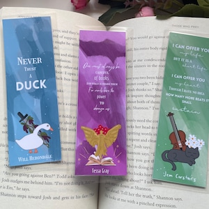 May include: Three bookmarks with different designs. The first bookmark is blue with a white duck and the text "Never Trust a Duck". The second bookmark is purple with a gold angel holding a book open and the text "One must always be careful of books and what is inside them. For words have the power to change us." The third bookmark is green with a black cat and a violin and the text "I can offer you my life but it is a short life. I can offer you my heart. Though I have no idea how many more beats it shall sustain."