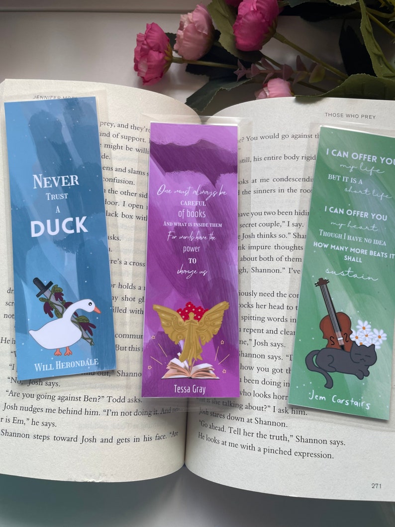 May include: Three bookmarks with different designs. The first bookmark is blue with a white duck and the text "Never Trust a Duck". The second bookmark is purple with a gold angel holding a book open and the text "One must always be careful of books and what is inside them. For words have the power to change us." The third bookmark is green with a black cat and a violin and the text "I can offer you my life but it is a short life. I can offer you my heart. Though I have no idea how many more beats it shall sustain."