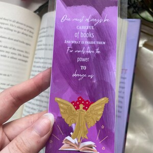 May include: Purple bookmark with a quote about books and an illustration of an angel with wings and a book. The quote reads "One must always be careful of books and what is inside them. For words have the power to change us." The bookmark is signed "Tessa Gray".