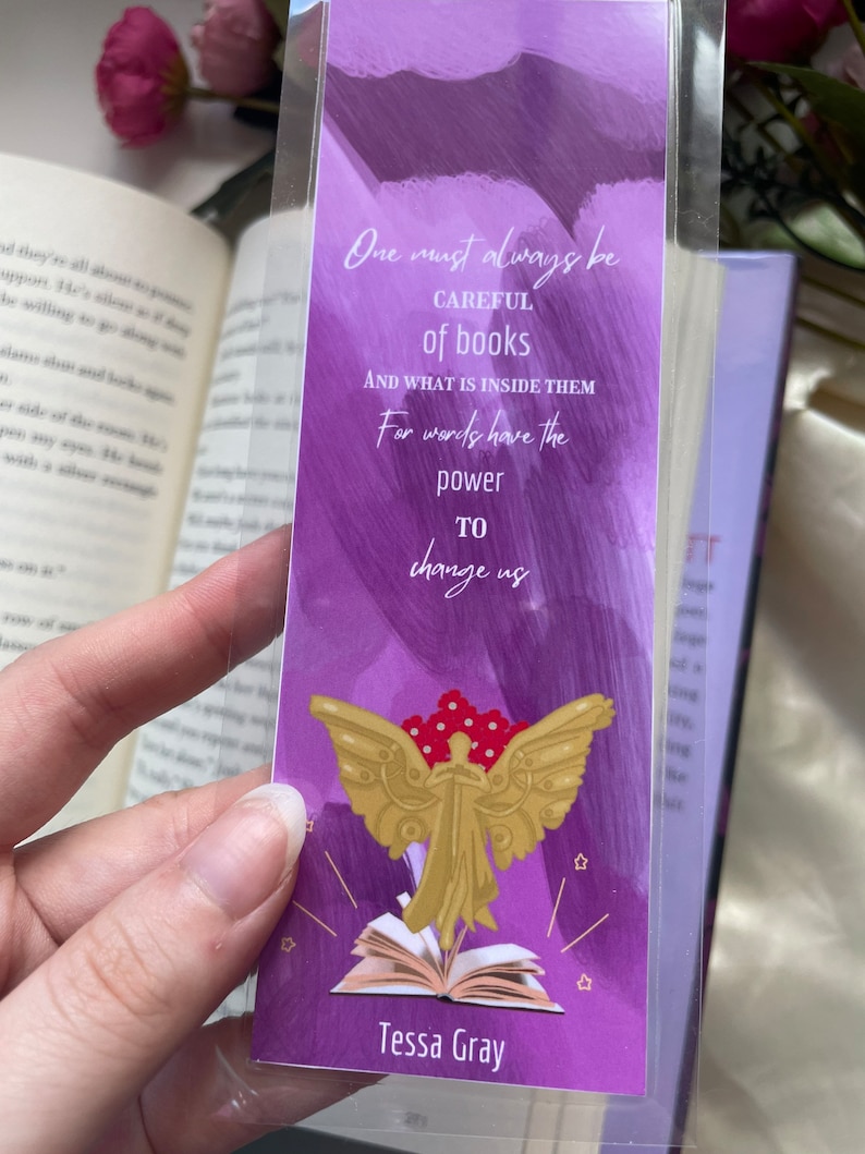 May include: Purple bookmark with a quote about books and an illustration of an angel with wings and a book. The quote reads "One must always be careful of books and what is inside them. For words have the power to change us." The bookmark is signed "Tessa Gray".