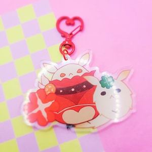 Puede incluir: Un llavero de acrílico transparente con tres personajes de dibujos animados. Los personajes son blancos con detalles rojos, naranjas y verdes. El llavero tiene un cierre en forma de corazón rojo.