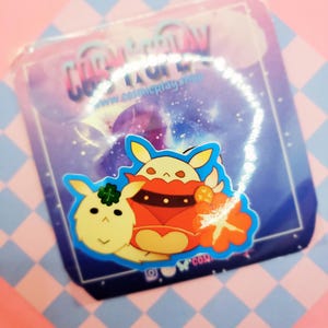 Puede incluir: Una pegatina con dos personajes de dibujos animados. El personaje de la izquierda es una criatura redonda, peluda y de color amarillo claro con un trébol en la cabeza. El personaje de la derecha es una criatura redonda, peluda y roja con un collar negro y una rodaja de naranja amarilla. La pegatina está sobre un fondo azul con un borde blanco.