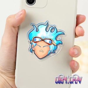 May include: A clear phone grip with a cartoon character design. The character has blue hair and goggles. The phone grip is attached to a light-coloured smartphone. The text "COSPLAY" is visible at the bottom of the phone.