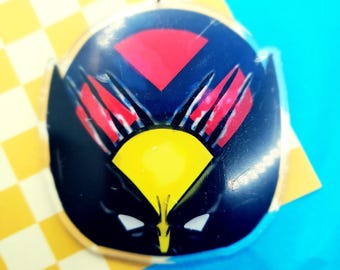 May include: A round, clear acrylic keychain with a black, yellow, and red graphic of Wolverine's face. The graphic features Wolverine's claws and a yellow mask.