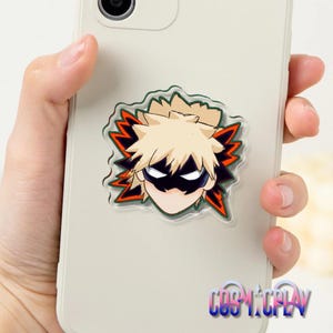 May include: A phone case featuring a cartoon character with spiky blonde hair, a black mask, and a determined expression. The character is set against a green and orange background. The phone case is held in a hand.