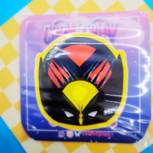 May include: A black and yellow sticker featuring a red and black Wolverine character design. The sticker has a white outline and the text "@owcosmicplay_" at the bottom.