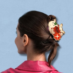 Puede incluir: Una pinza para el pelo dorada con un diseño rojo y blanco con dos conejos de dibujos animados. Los conejos sostienen una flor roja con un centro amarillo.