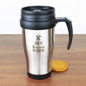 May include: Silver travel mug with a black lid and handle. The mug has a gold inscription that reads "DIY Expert DAVE" with a wrench and hammer symbol.