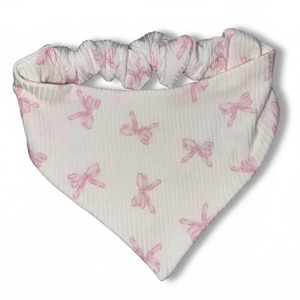 May include: White and pink baby bib with a bow pattern. The bib is made of a soft, ribbed fabric and features a pink bow print. The top of the bib has a gathered, elasticized band.