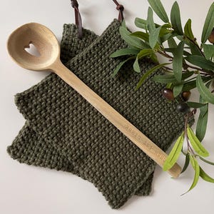 Pode incluir: Uma colher de pau com um recorte em forma de coração e o texto gravado "Kochen ist Liebe" repousa sobre dois panos de cozinha de crochê verde-oliva. Laços de couro castanho estão presos aos panos de cozinha. Ramo de oliveira com azeitonas no fundo.