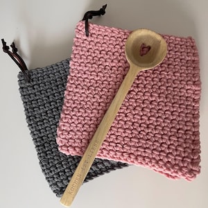 Könnte beinhalten: Ein Holzlöffel mit herzförmigem Ausschnitt liegt auf einem rosa gehäkelten Topflappen. Ein grauer gehäkelter Topflappen und ein Holzlöffel mit dem Text "KOCHEN IST LIEBE" sind ebenfalls im Bild. Beide Topflappen haben Lederschlaufen.