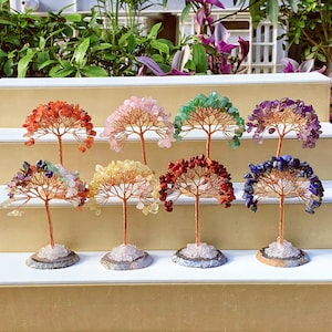 May include: Ten small wire trees decorated with colourful gemstones. Each tree is on a white base and arranged in a row on a wooden shelf.