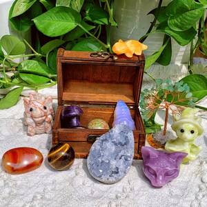 Pode incluir: Um ba&uacute; de madeira cheio de v&aacute;rios cristais e estatuetas. O ba&uacute; est&aacute; aberto e os itens dentro incluem um cogumelo roxo, um cristal azul, um cristal verde, uma estatueta amarela, uma estatueta branca e uma estatueta roxa.