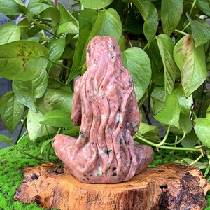 May include: A pink and green stone sculpture of a woman sitting with her legs crossed and her arms resting on her lap. The sculpture is carved in a detailed style, with flowing lines and a natural, organic look.