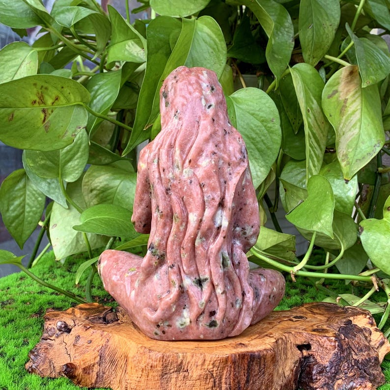May include: A pink and green stone sculpture of a woman sitting with her legs crossed and her arms resting on her lap. The sculpture is carved in a detailed style, with flowing lines and a natural, organic look.