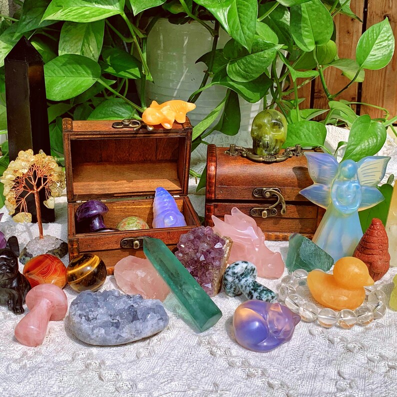Pode incluir: Uma cole&ccedil;&atilde;o de cristais, pedras e estatuetas coloridas dispostas sobre uma toalha de mesa branca. Os itens incluem um quartzo rosa, uma aventurina verde, uma ametista roxa, um quartzo transparente, um lapis-laz&uacute;li azul, um olho de tigre amarelo, uma &aacute;gata marrom, uma estatueta de anjo branca, uma estatueta de pato amarela e um ba&uacute; de madeira.