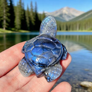 Puede incluir: Una figura de tortuga de piedra preciosa tallada con un brillo iridiscente azul y gris. La tortuga se sostiene en una mano, con un lago y montañas al fondo. El caparazón de la tortuga tiene un patrón geométrico.