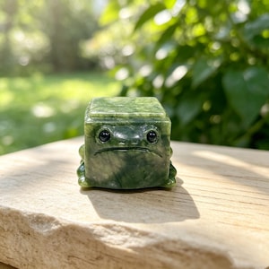 May include: A small, cube-shaped figurine carved from mottled dark green stone, featuring a stylised frog face with two smooth, black, circular inlaid eyes and a simple horizontal line carved to represent a mouth on the front surface.