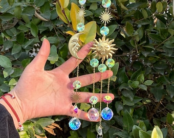 May include: A celestial-themed suncatcher with a gold-colored sun and moon pendant, adorned with iridescent crystal beads and star accents. The suncatcher is designed to hang, adding a decorative touch to any space.
