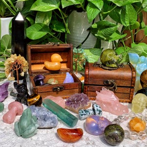 Pode incluir: Uma cole&ccedil;&atilde;o de cristais, pedras e estatuetas dispostas sobre uma superf&iacute;cie branca. Os itens incluem uma caveira verde, um pato de borracha amarelo, um sapo verde, um drag&atilde;o de quartzo rosa, um aglomerado de ametista roxo, uma aventurina verde, um quartzo claro, um olho de tigre, um cogumelo de quartzo rosa, uma esfera de aventurina verde, uma esfera de quartzo claro, uma torre de obsidiana preta e uma pequena &aacute;rvore feita de fio de cobre e contas.